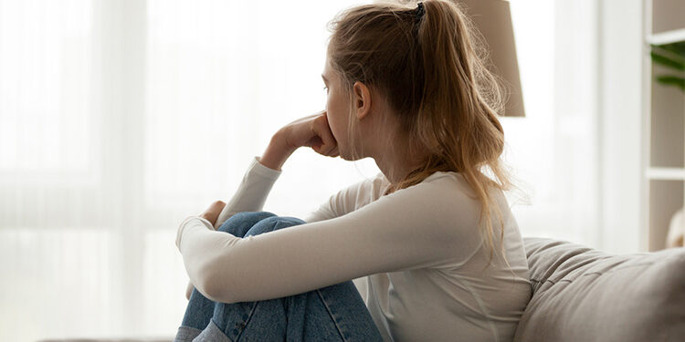 Side view young woman looking away at window sitting on couch at home. Frustrated confused female feels unhappy problem in personal life quarrel break up with boyfriend or unexpected pregnancy concept