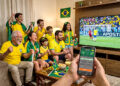 Pessoa segurando um smartphone na sala de casa exibindo gráficos e propagandas de apostas esportivas de futebol na tela.