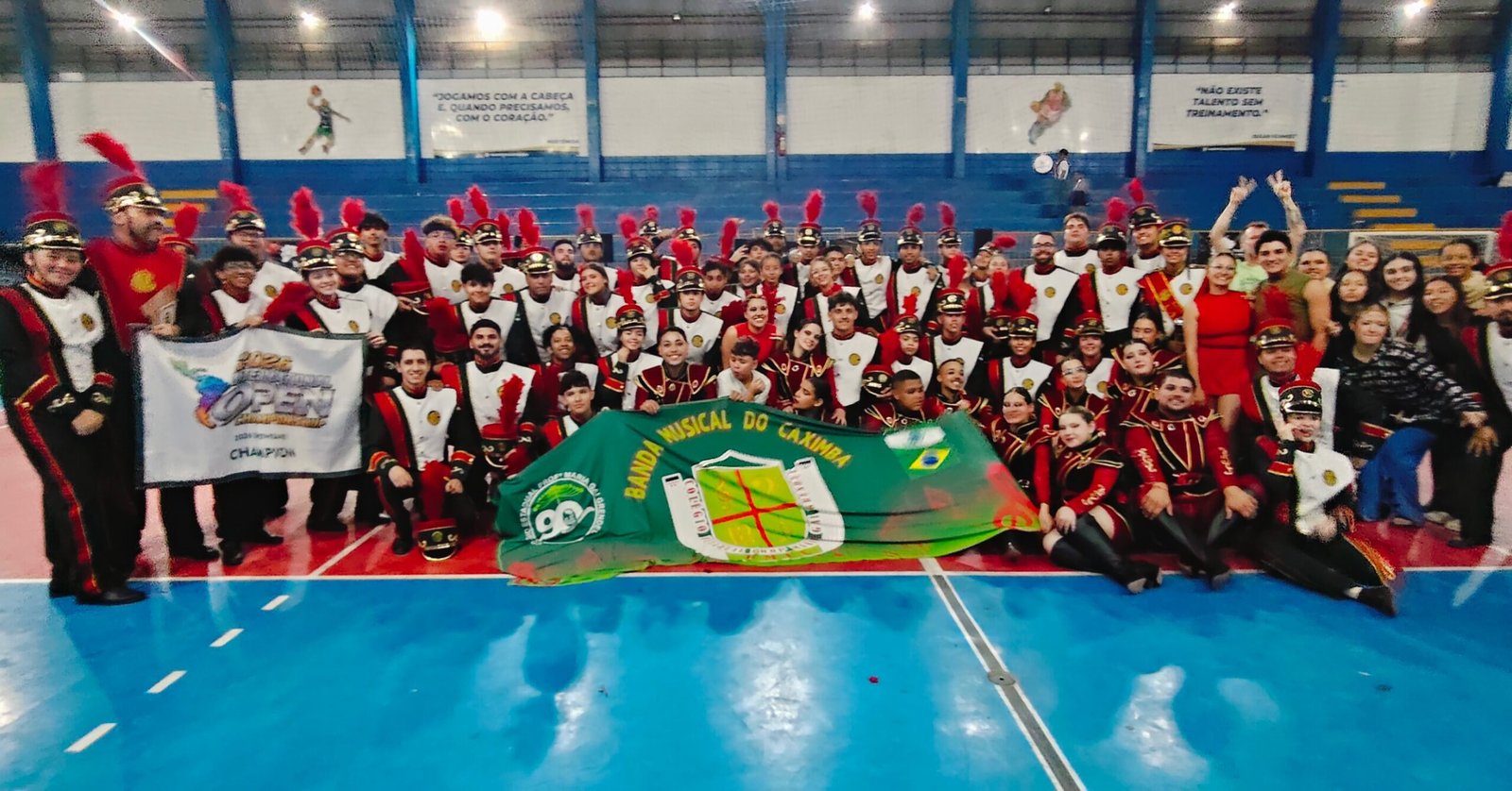 Banda Musical do Caximba reunida em um ginásio com bandeira e faixa de campeã.
