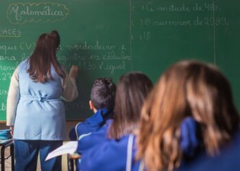 Professora escreve no quadro diante de alunos em sala de aula.