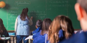 Professora escreve no quadro diante de alunos em sala de aula.