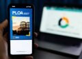 Mão segura celular com a capa da PLOA 2027 de Curitiba, diante de um notebook.