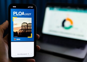 Mão segura celular com a capa da PLOA 2027 de Curitiba, diante de um notebook.
