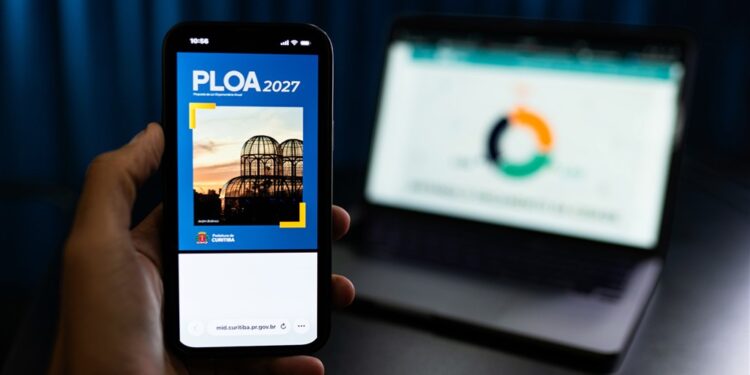 Mão segura celular com a capa da PLOA 2027 de Curitiba, diante de um notebook.