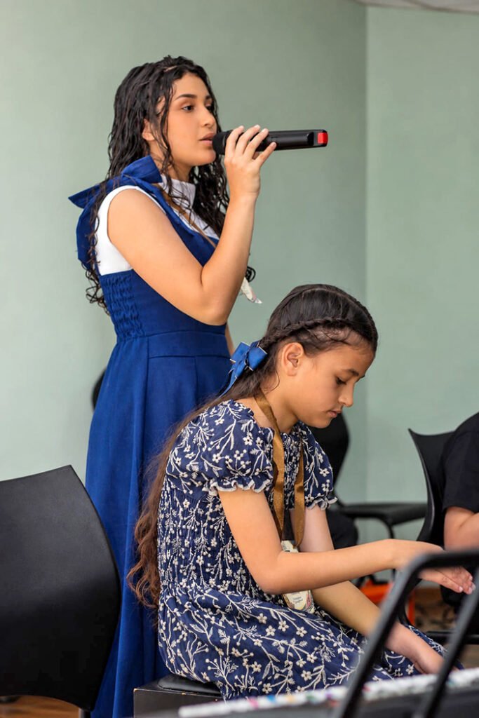 Aluna canta ao microfone enquanto outra aluna toca teclado no recital.
